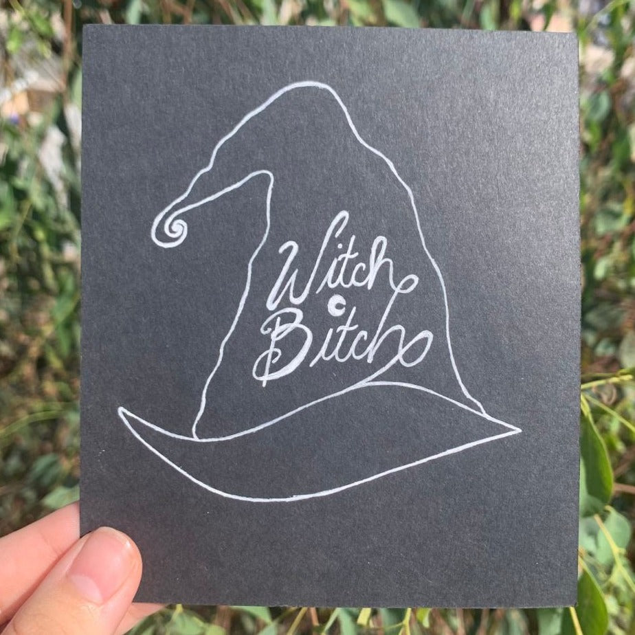 Witch Bitch original drawing on black paper with white pen featuring a witch's hat with a swirl at the end and cursive text calligraphy lettering topped with a crescent moon 